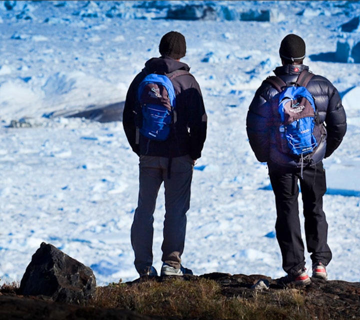 A pair of men on a bachelor destination trip to Patagonia wearing backpacks as they stand on a cliff overlooking glaciers